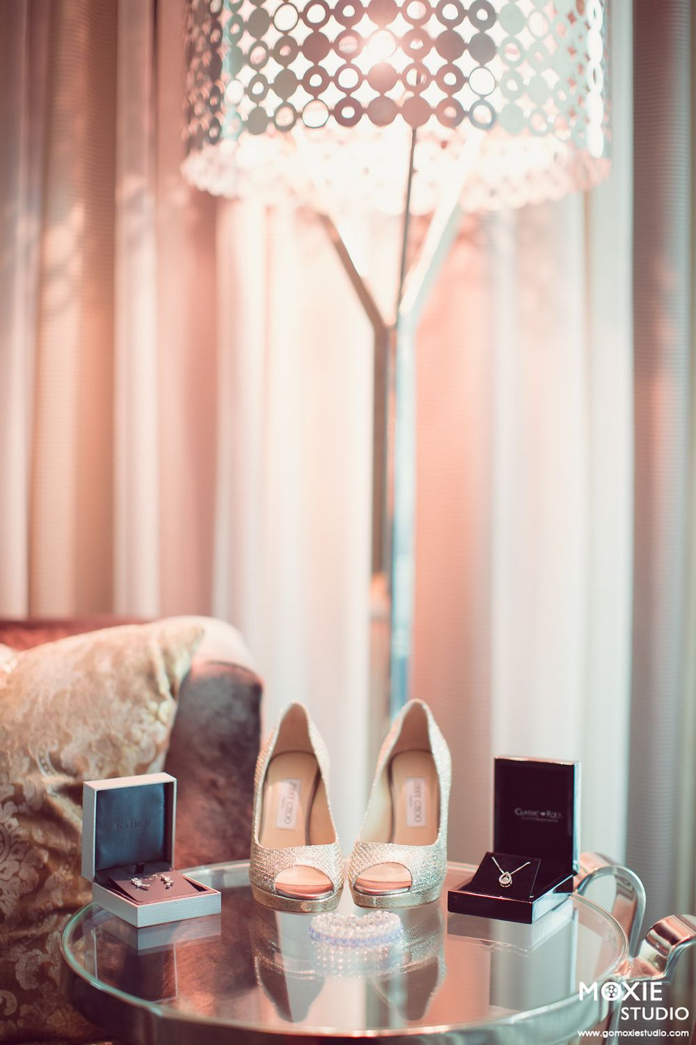 Wedding accessories including blush pink heels, jewelry boxes and a decorative lamp on a round mirrored table.
