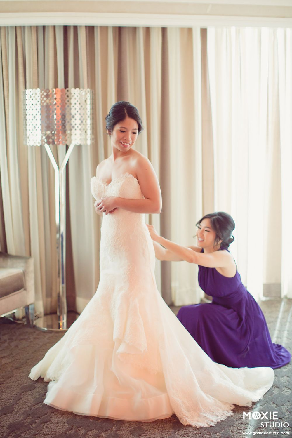 Bridesmaid in purple dress helping adjust the bride's flowing white wedding gown.