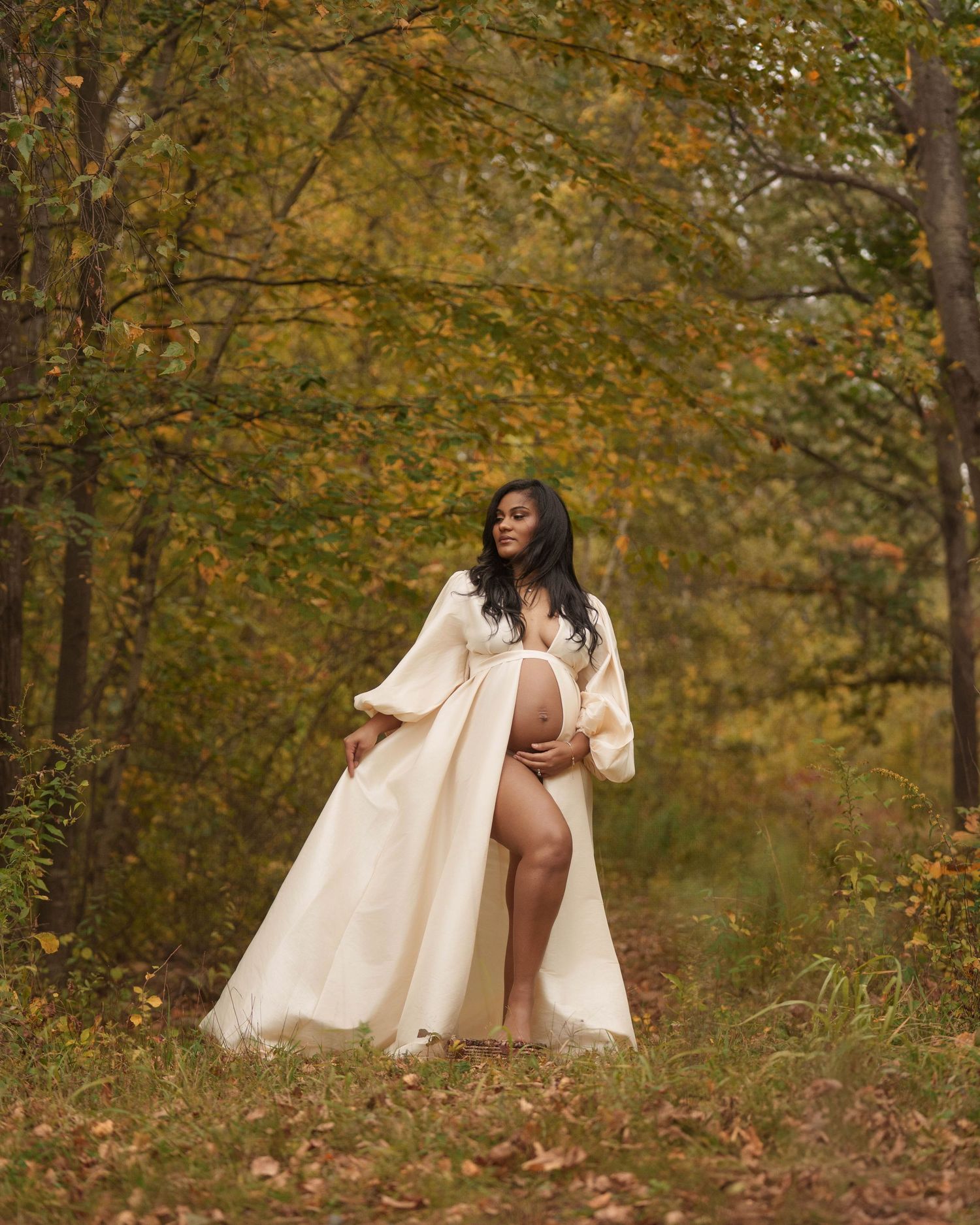 Artistic maternity portrait in a cream robe against autumn foliage with warm golden tones in a natural outdoor setting.