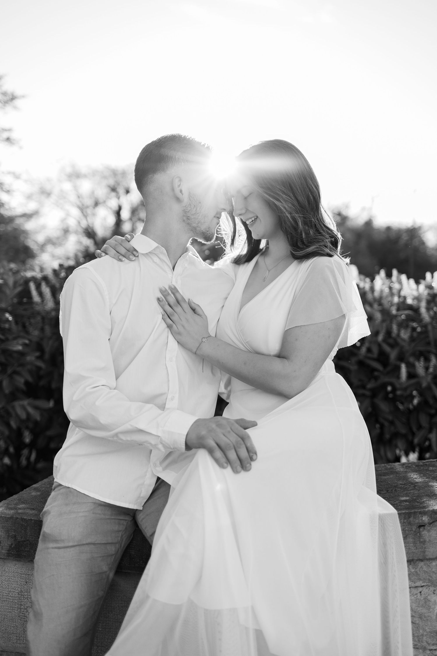 Black and white photo of a couple in white outfits sharing a romantic moment outdoors with sunlight glowing between them.