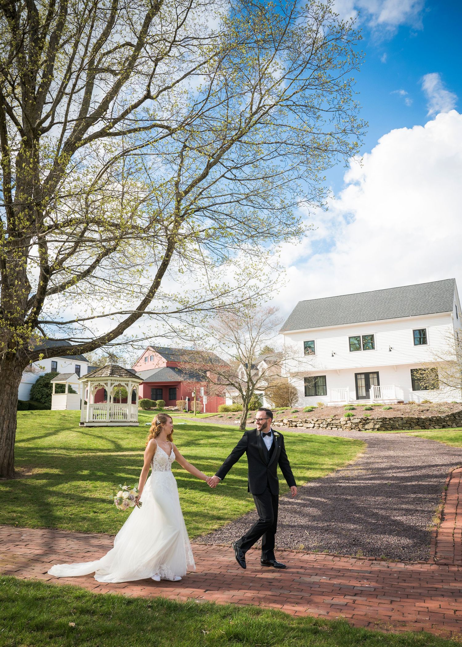 A couple in wedding attire walks along a brick path surrounded by green lawns and white buildings on a sunny day.
