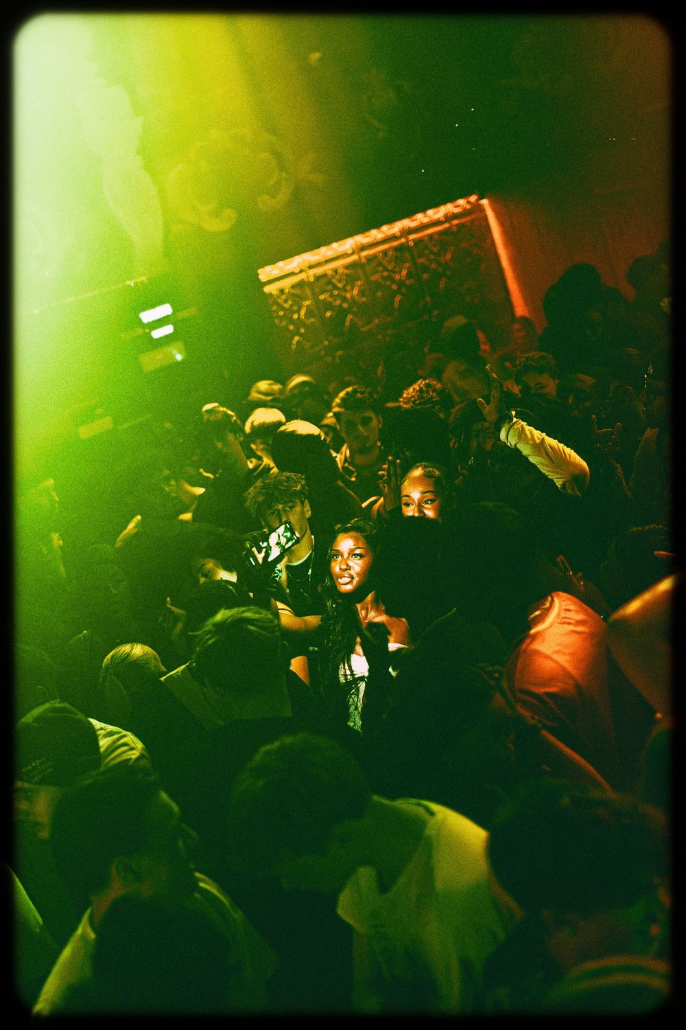 Crowded nightclub scene bathed in green lighting shows silhouettes of people dancing and socializing on the dance floor.