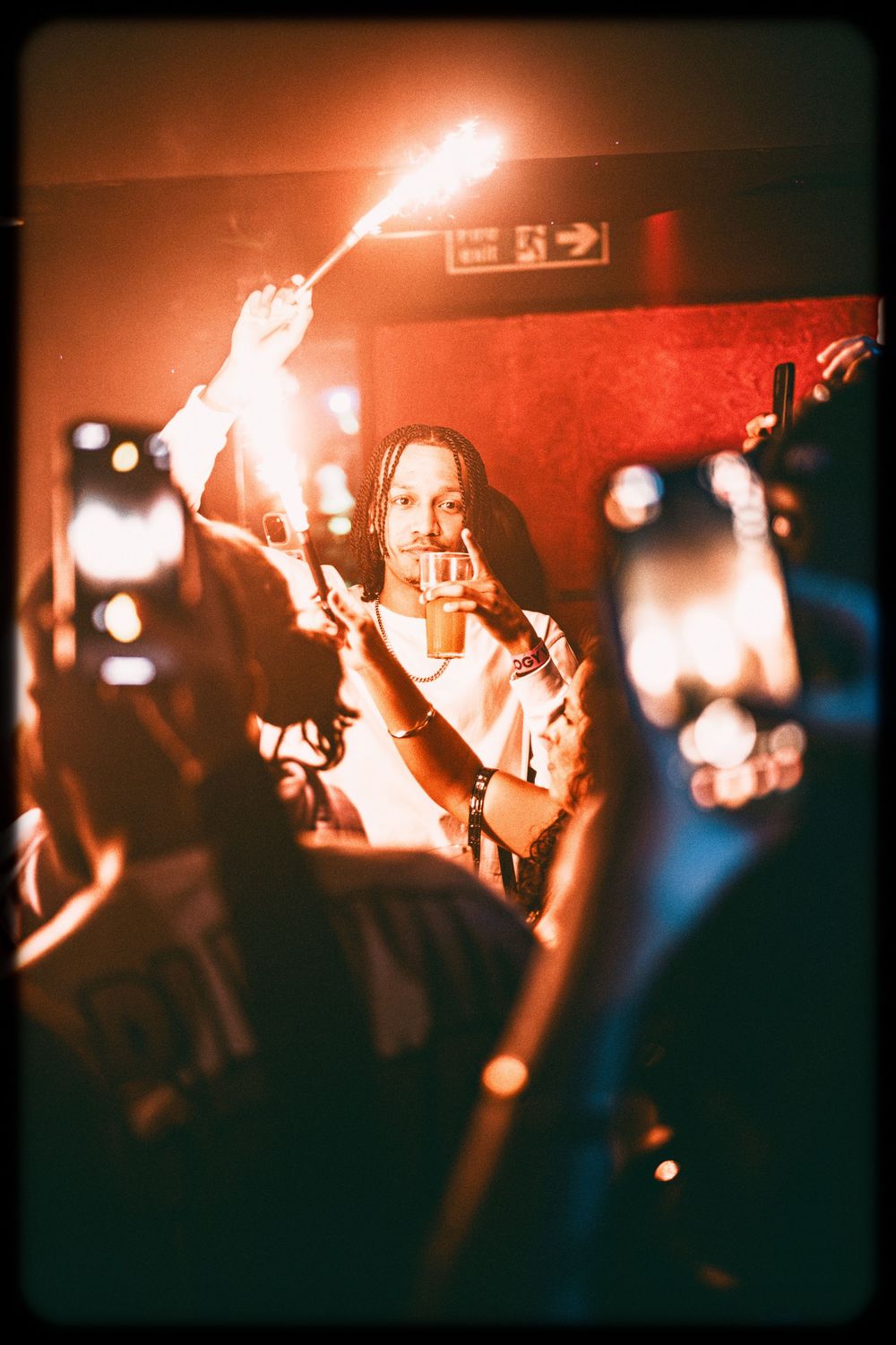 A person holds a bright flare in a dark crowd during a nighttime gathering with red lighting and silhouettes visible.