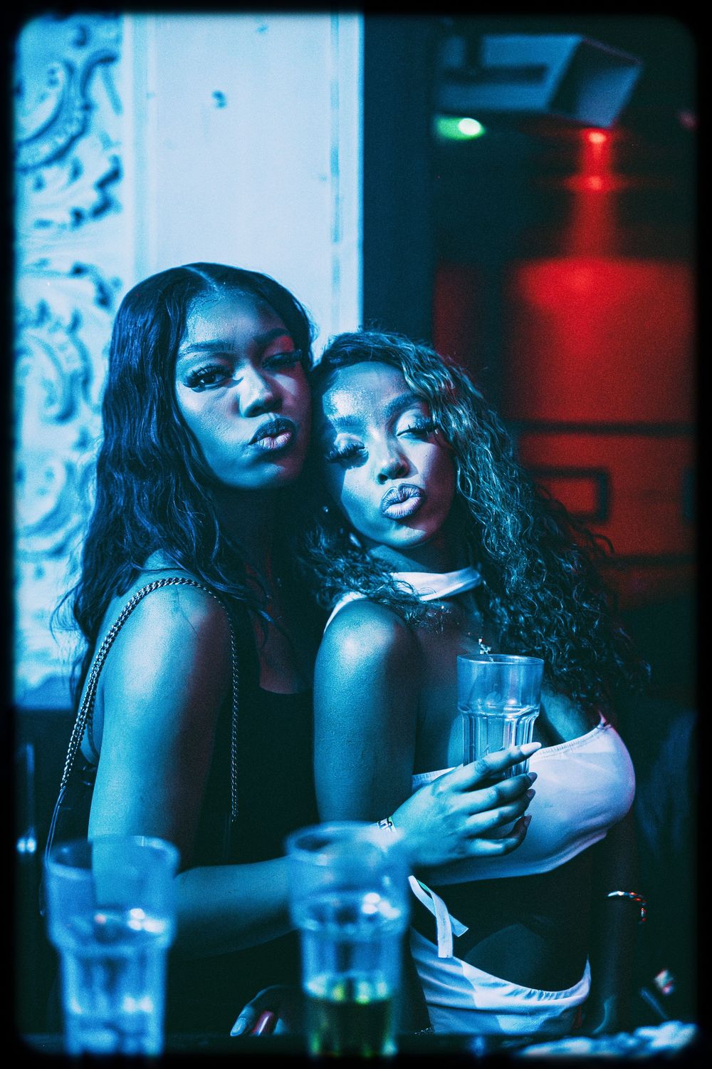 Two people pose together at a nightclub with drinks in a moody blue and red lit atmosphere.