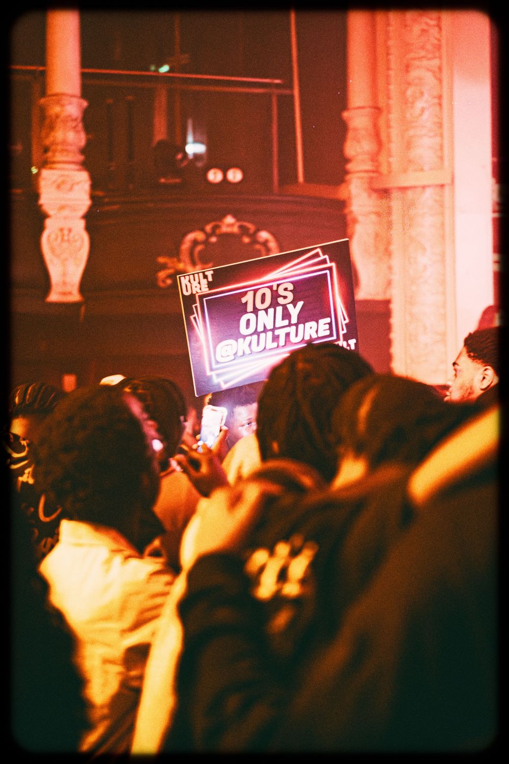 A crowd holds illuminated signs at a nighttime event with an orange-tinted atmospheric glow.