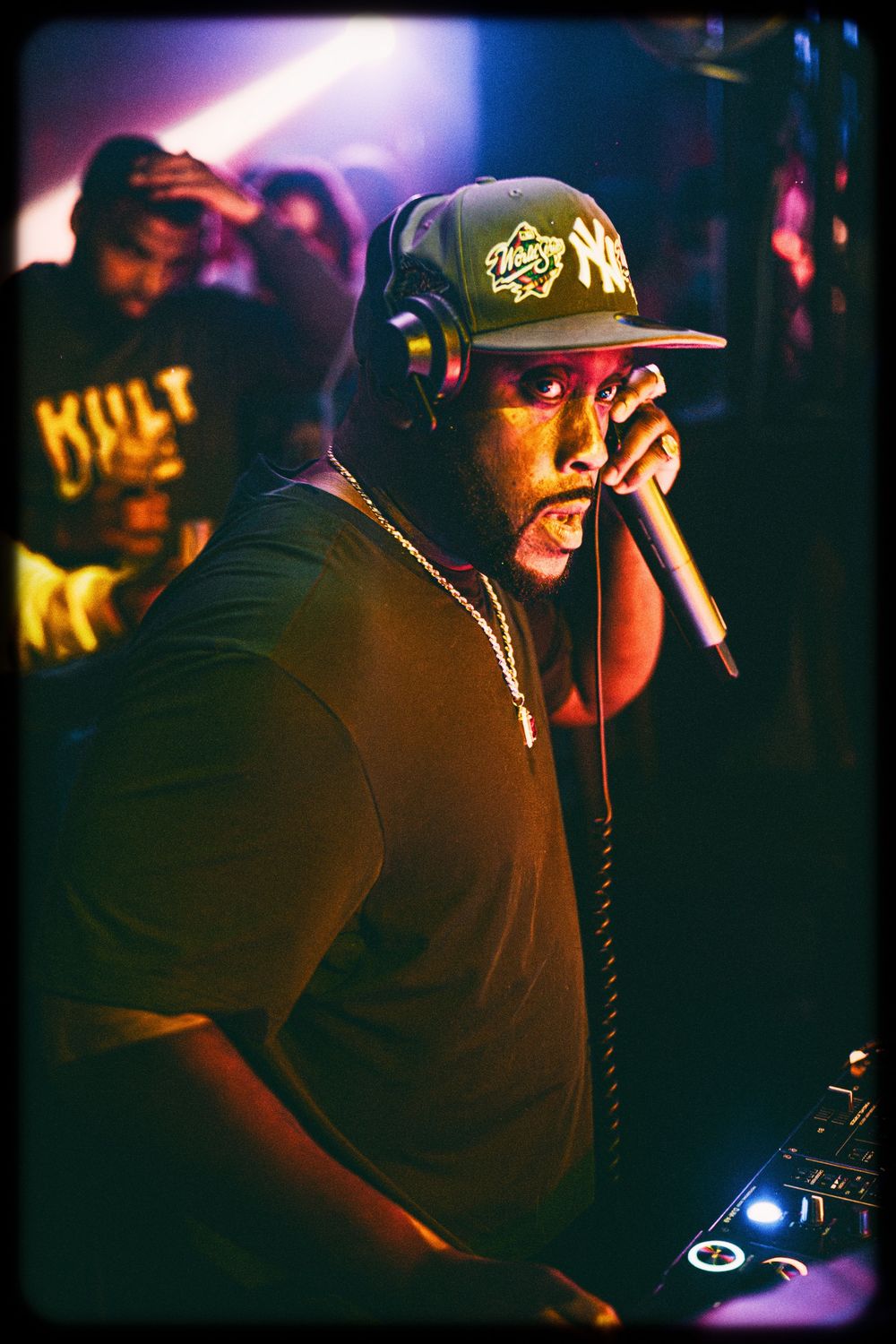 Hip hop performer wearing headphones and a snapback cap performs on stage in dramatic lighting.