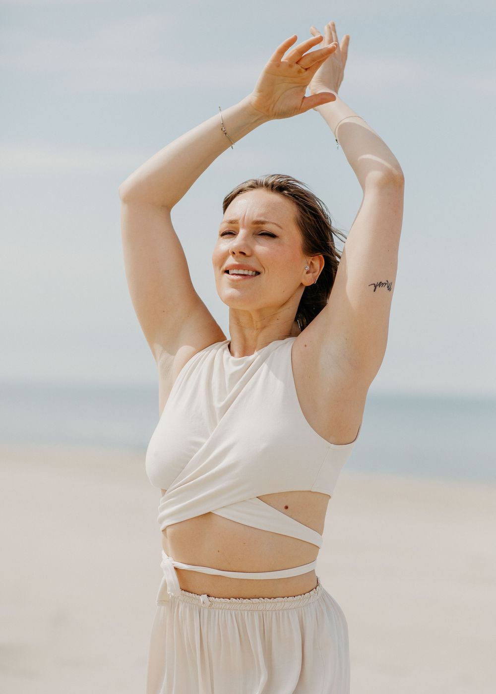 Woman in white beach outfit smiles with arms raised overhead against a cloudy sky.
