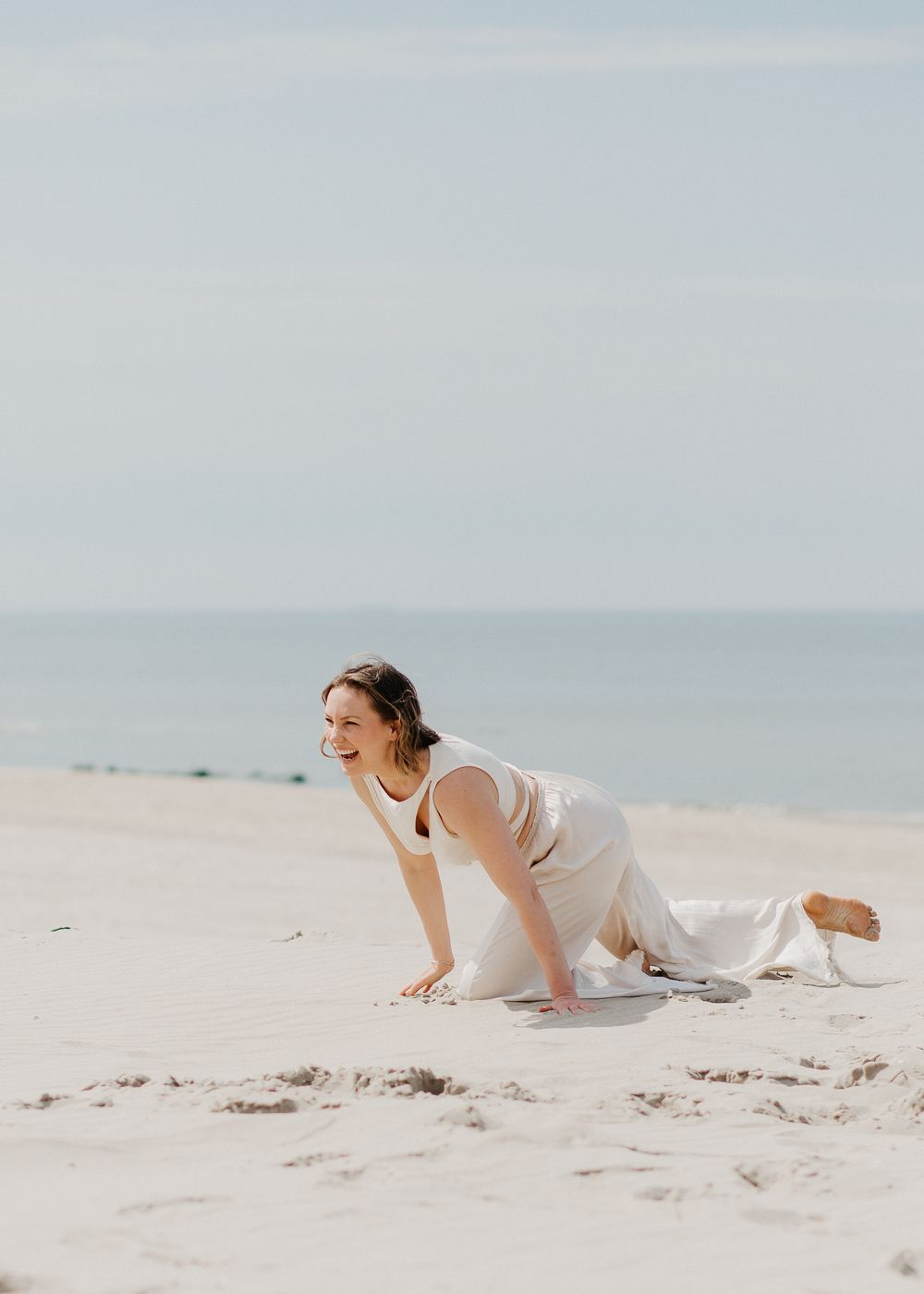 Woman in white outfit laughs while crawling playfully on a sandy beach shoreline.