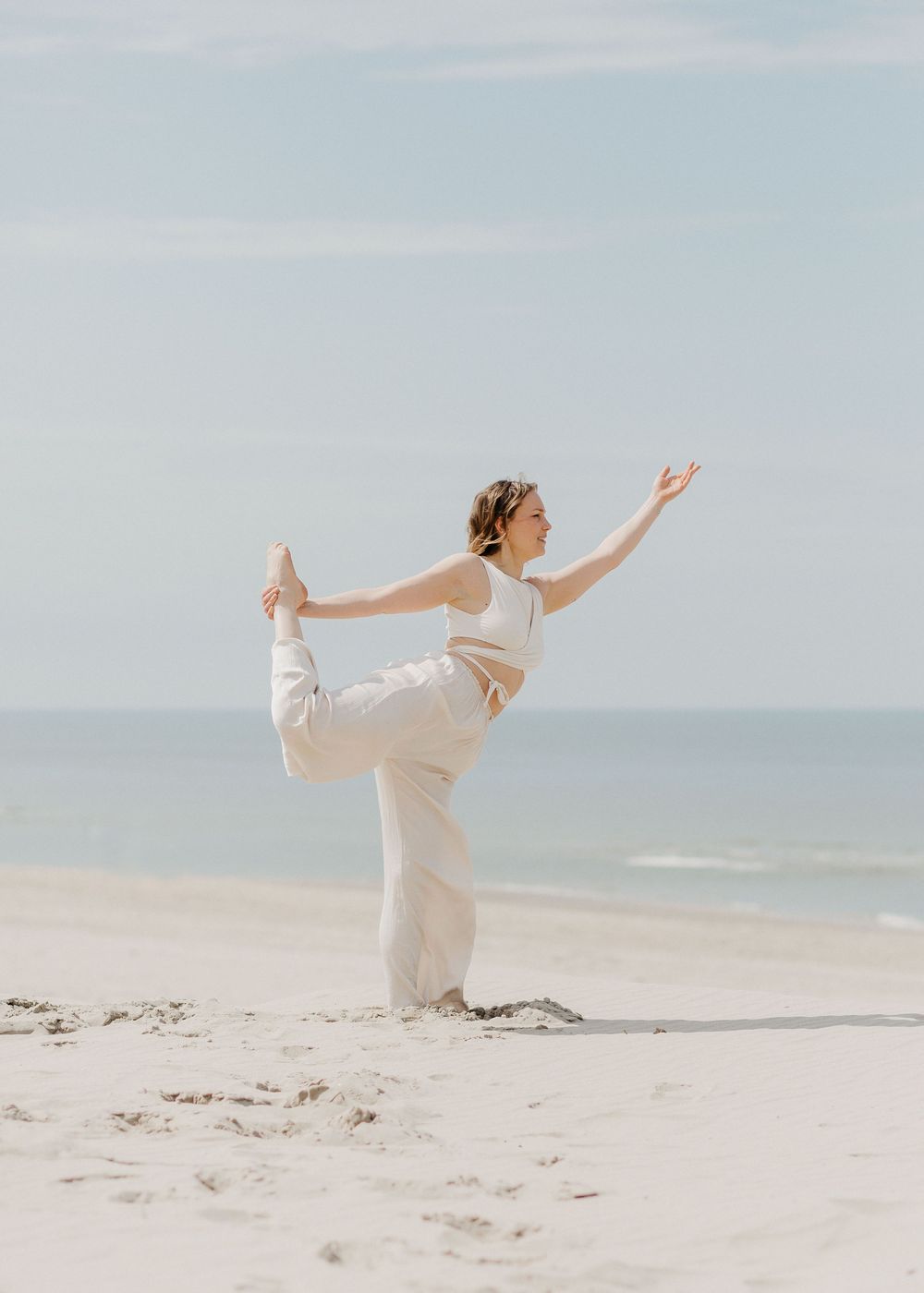 Woman in white outfit performs a yoga dancer pose on a sandy beach by the ocean.