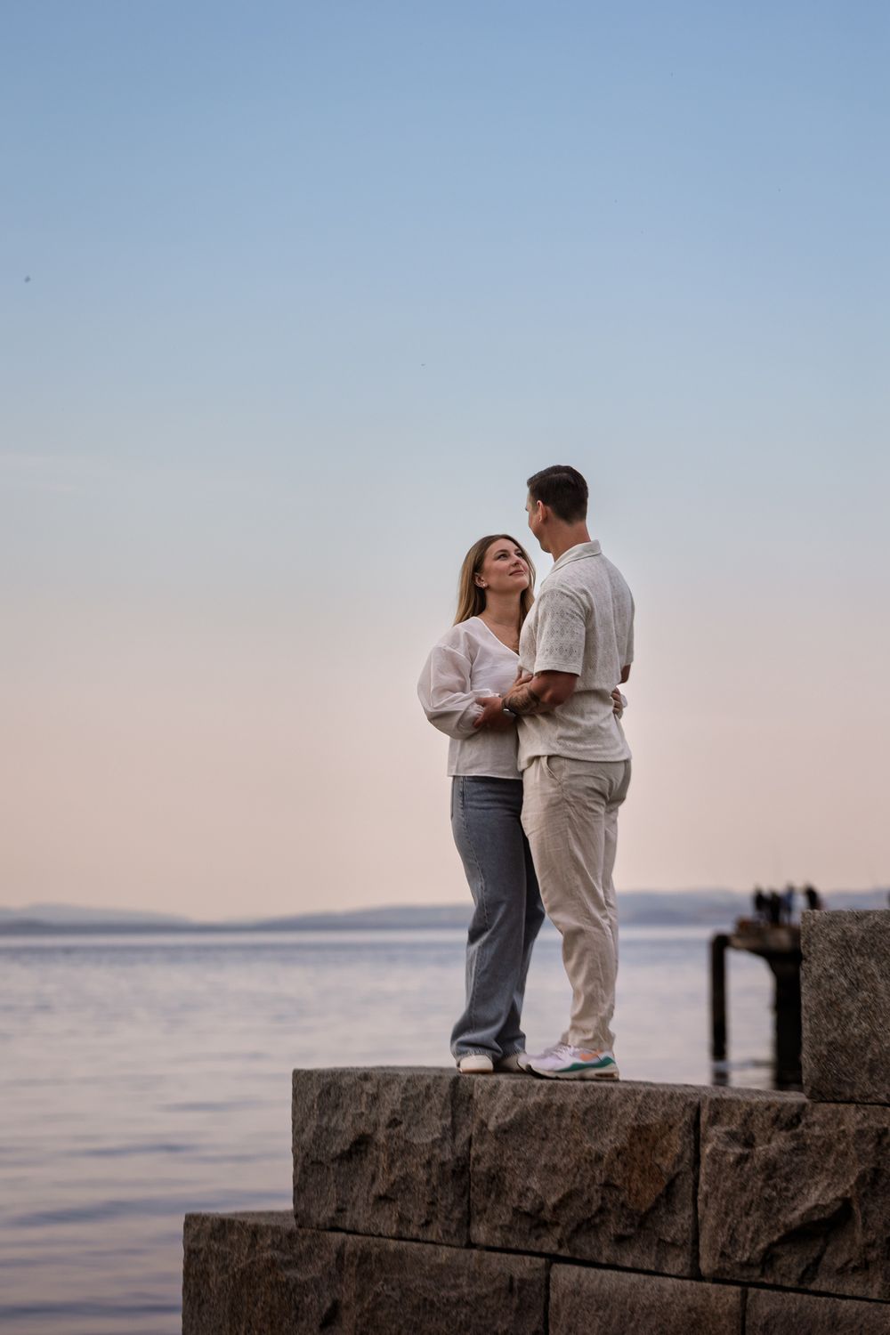 A romantic couple embraces on a stone wall overlooking a serene ocean view at sunset.