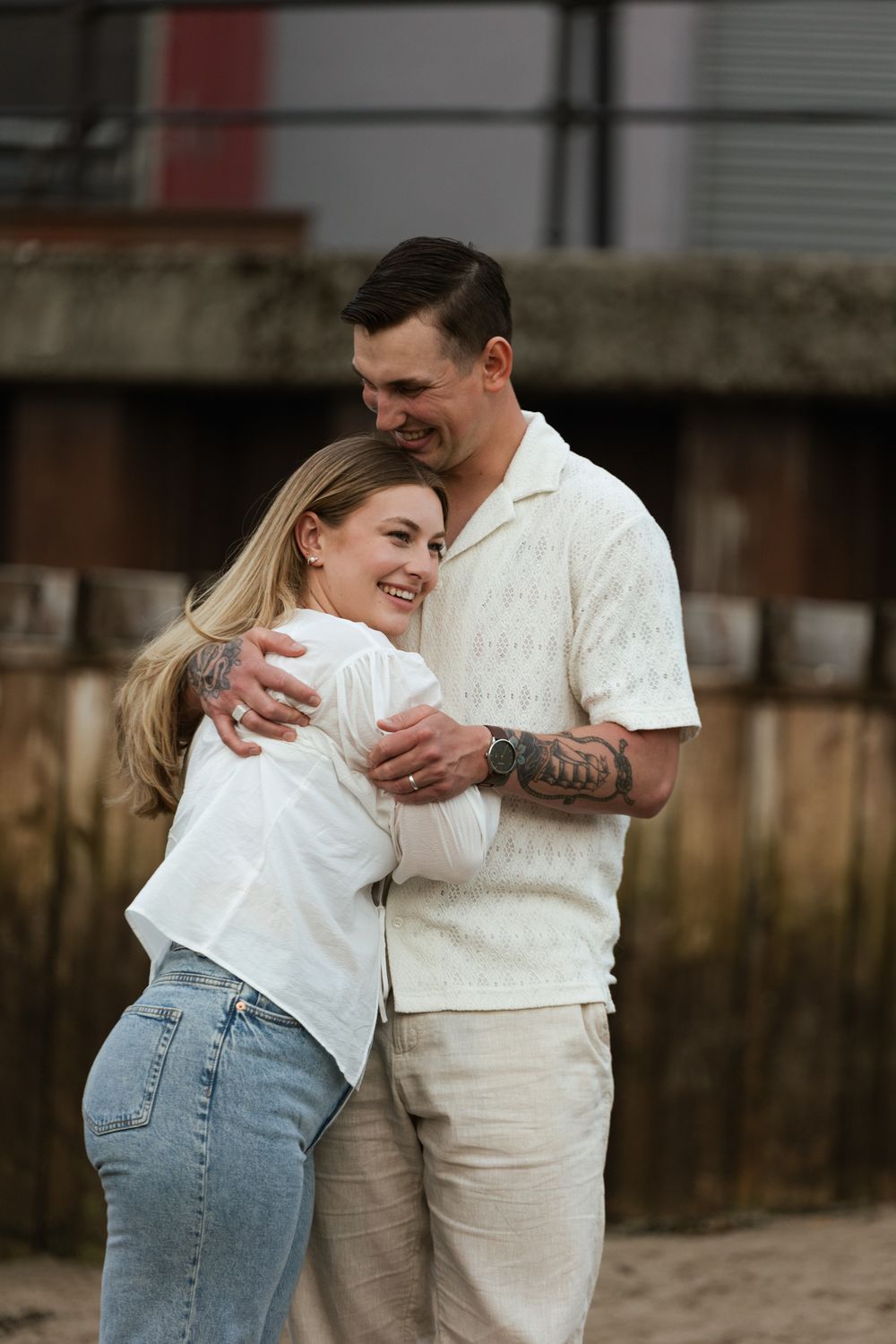 A couple in white shirts and denim embraces lovingly on a sandy beach during a romantic photoshoot.