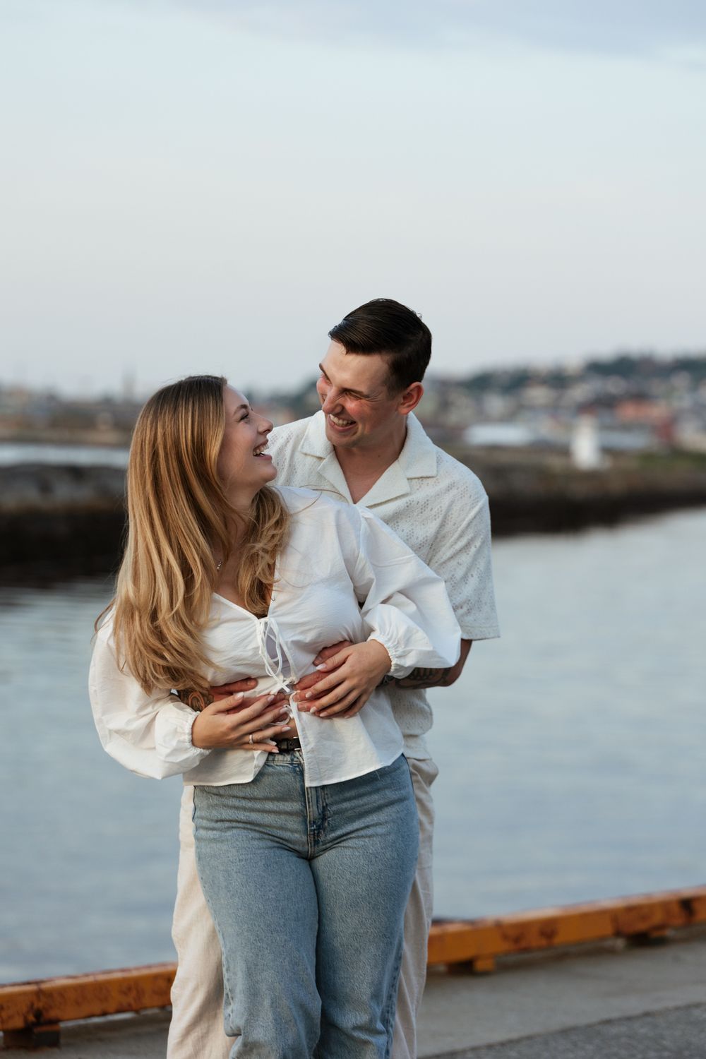 A romantic couple shares a tender moment while standing together on a waterfront pier at sunset.
