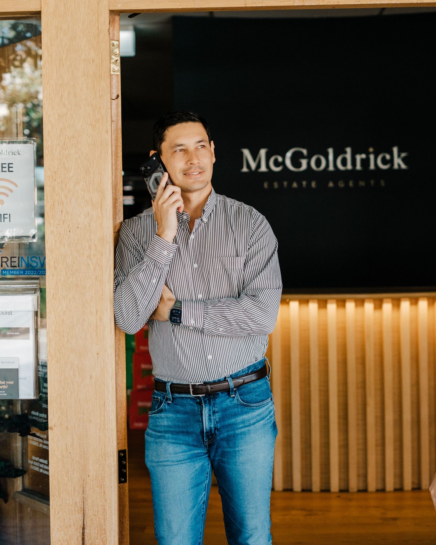 Person in striped button-down shirt and blue jeans poses casually against wooden wall in office setting.