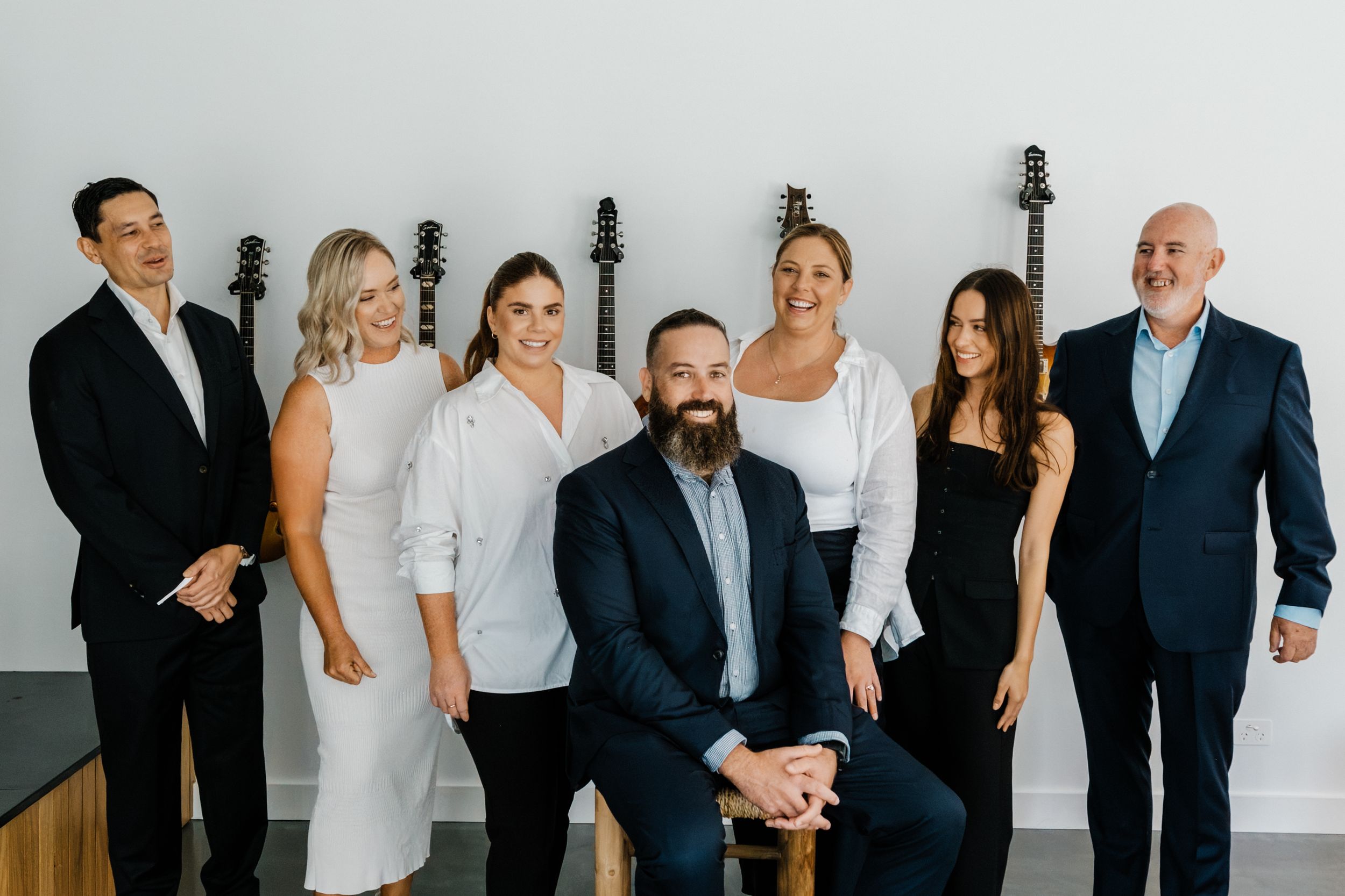 A professional real estate team of seven smiling adults posing together in a modern white studio with guitars on the wall.