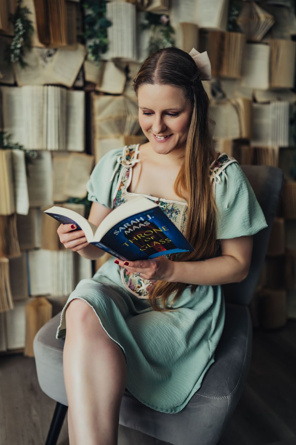 A smiling woman in a mint dress reads Sarah J. Maas Throne of Glass book seated in a chair before a wall of open books.