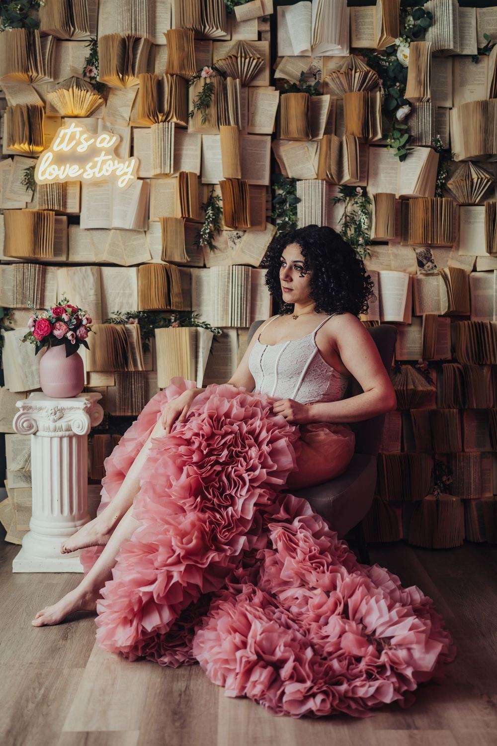 Curly haired woman in white corset sitting in a voluminous pink ruffled skirt by a wall of books.
