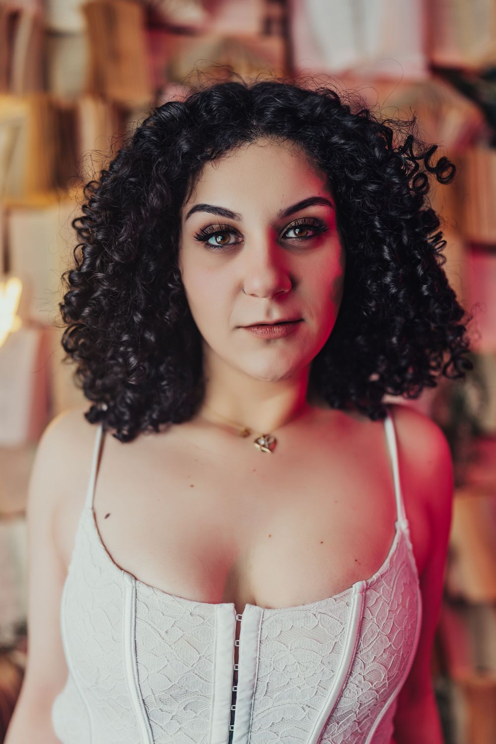 Woman with curly black hair and blue eyes wearing a white corset top against a wooden backdrop.