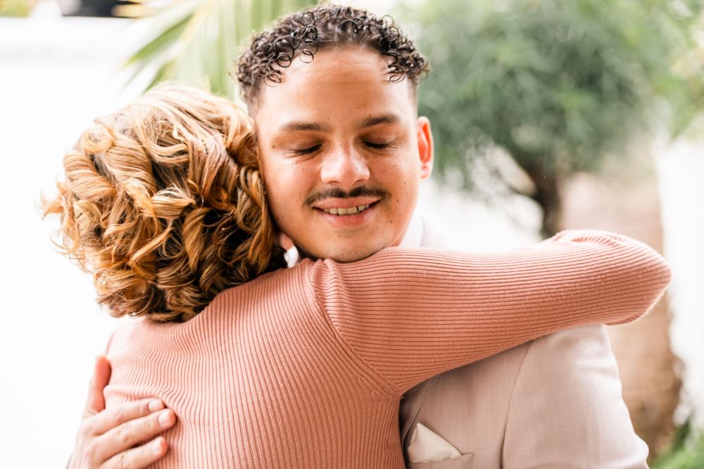 Two people share a warm embrace outdoors in natural lighting with a soft pink sweater visible.