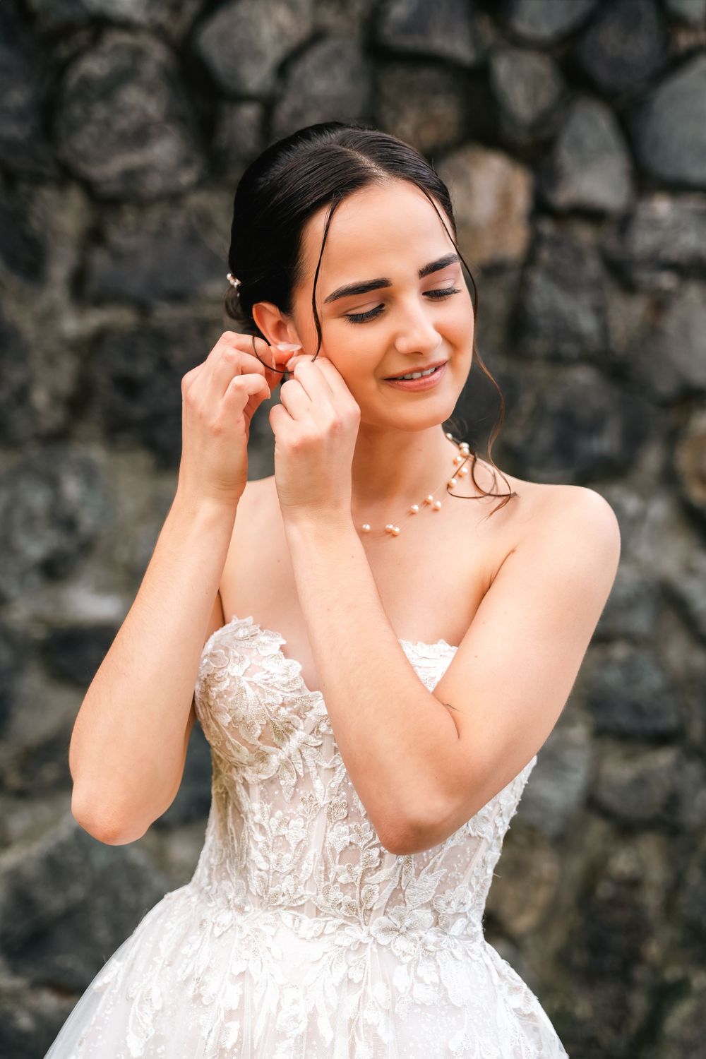 A person in a white lace wedding dress poses expressively against a stone wall background.