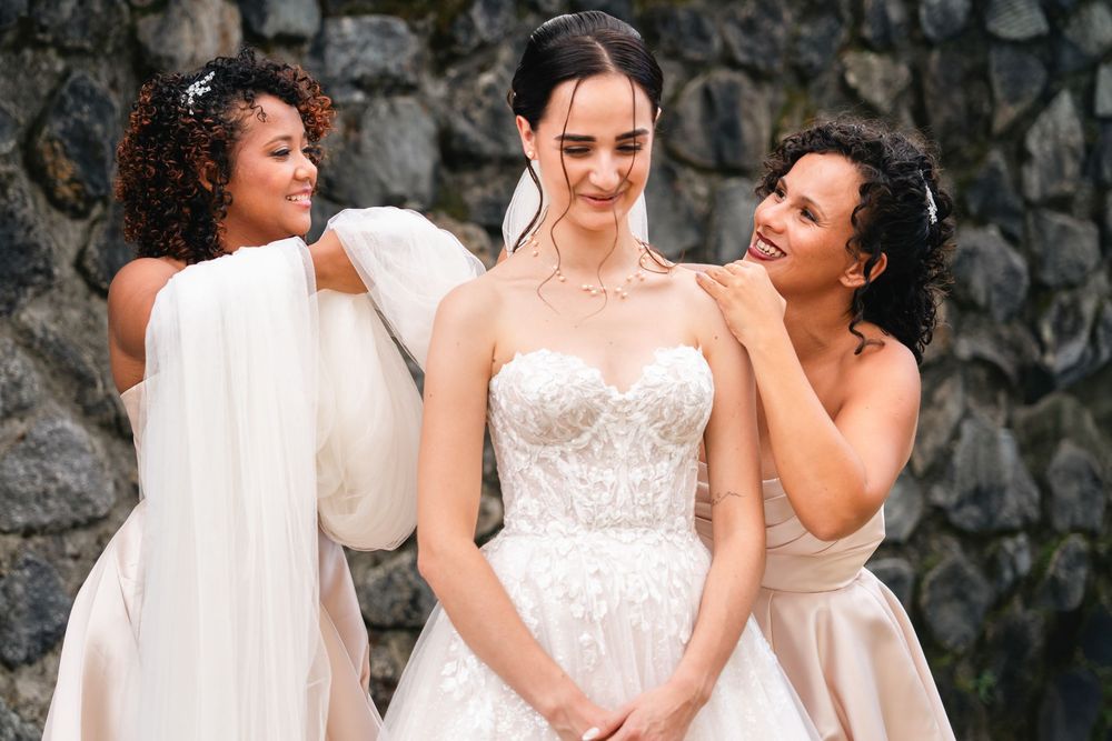 Three women in white dresses laugh joyfully together against a stone wall background during a formal event.