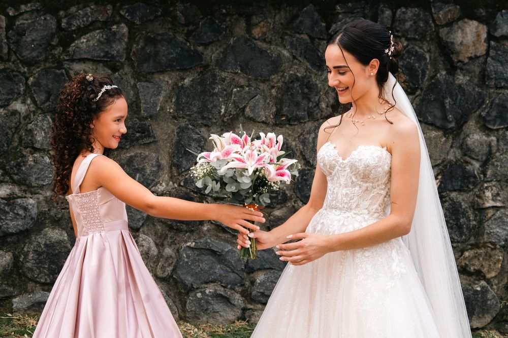 A young bridesmaid in pink dress hands a pink and white bouquet to a bride wearing a lace wedding gown against stone wall.
