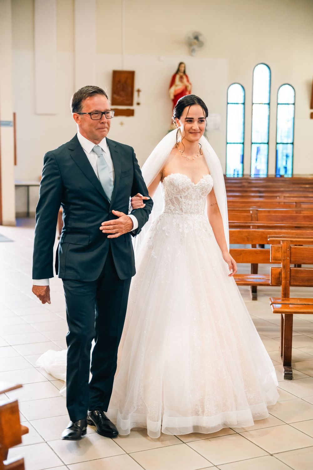 A bride in a white lace ballgown walks down the church aisle with an escort in a dark suit.