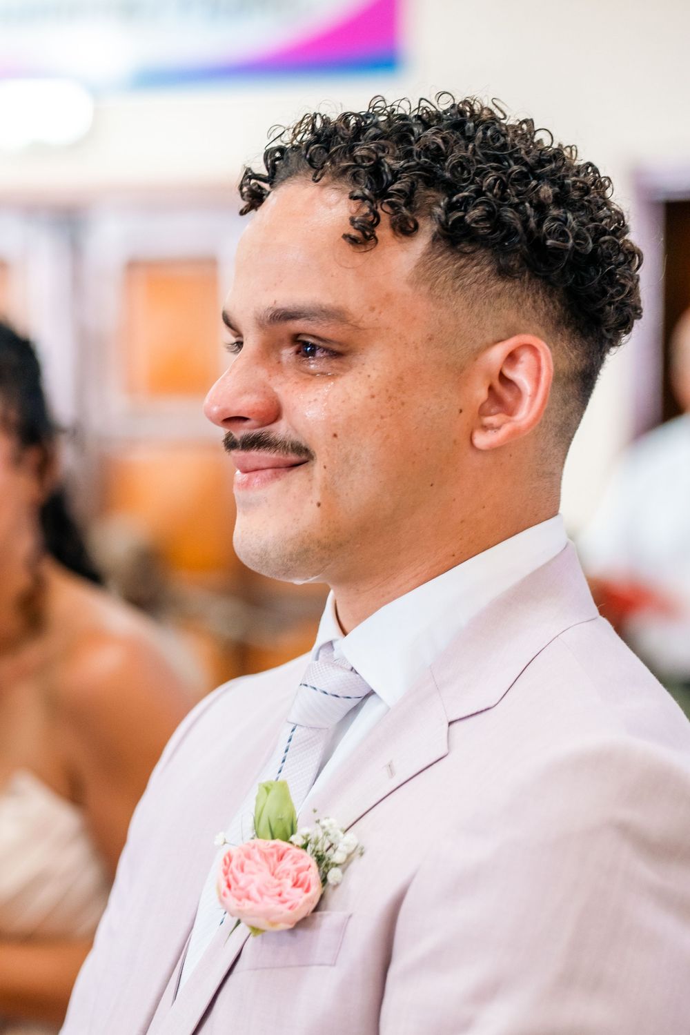 Close-up profile view of a groom wearing a light beige suit with a pink rose boutonniere at a wedding ceremony.
