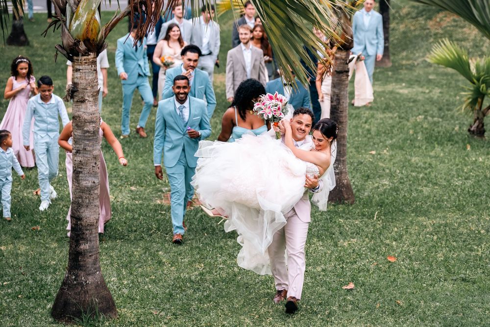 Wedding guests in light blue and white attire gather on a tropical lawn with palm trees.