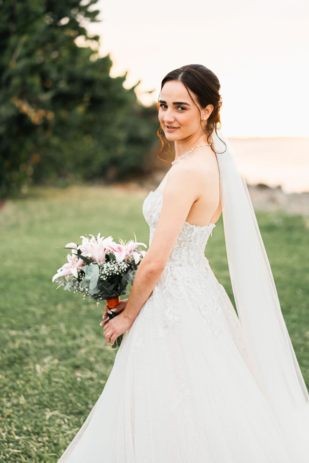 A bride in a white lace wedding dress and flowing veil poses outdoors during sunset in a grassy field.