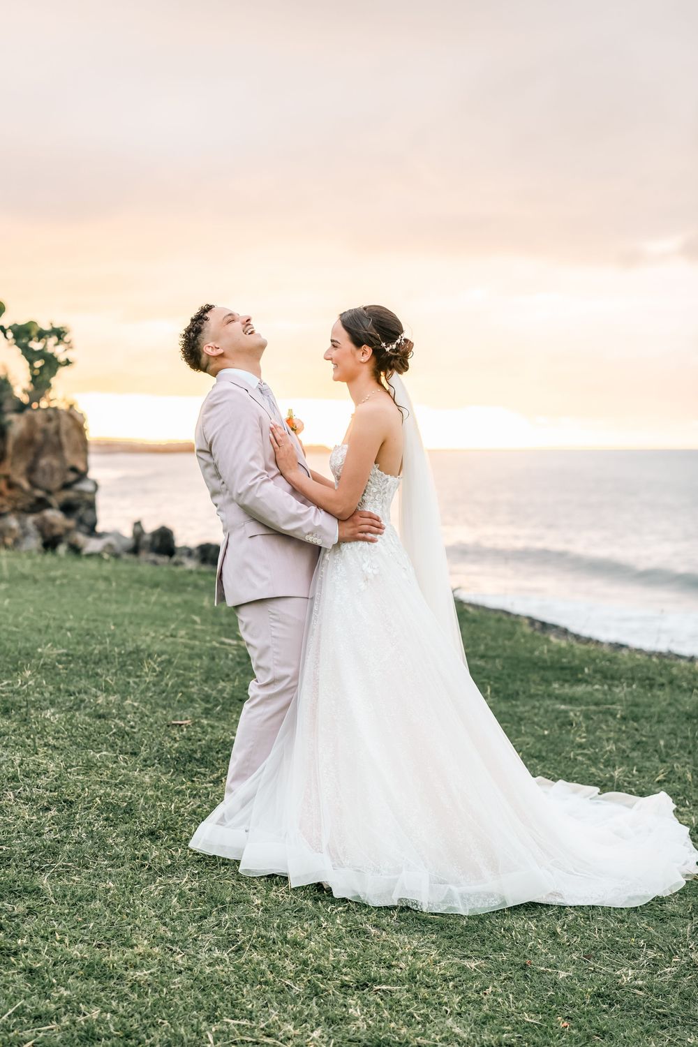 A romantic wedding couple shares an intimate moment on a grassy oceanside cliff during sunset.