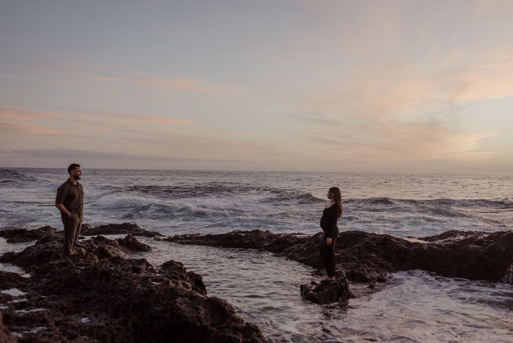 Two silhouettes stand on rocky coastline during sunset as waves crash against the shore.