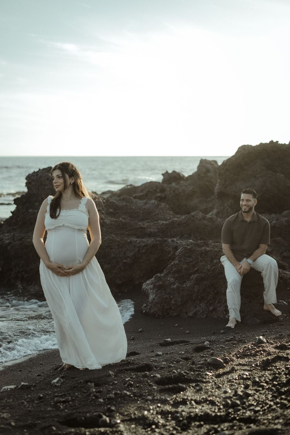 A couple poses on a rocky beach at sunset, with a pregnant woman in a flowing white dress standing by the water.