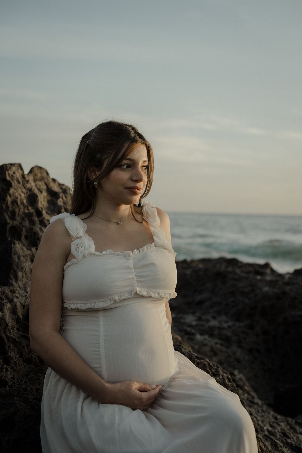 A woman in a white dress sits thoughtfully on coastal rocks at sunset while waves crash in the background.