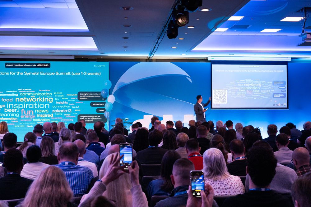 Conference attendees gather in a blue-lit auditorium watching presentations on large projection screens.