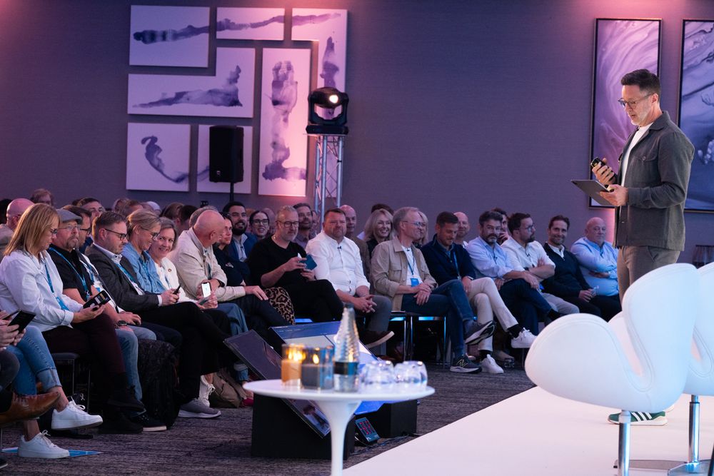 A speaker presents to an attentive audience in a dimly lit room with purple lighting and wall decorations.