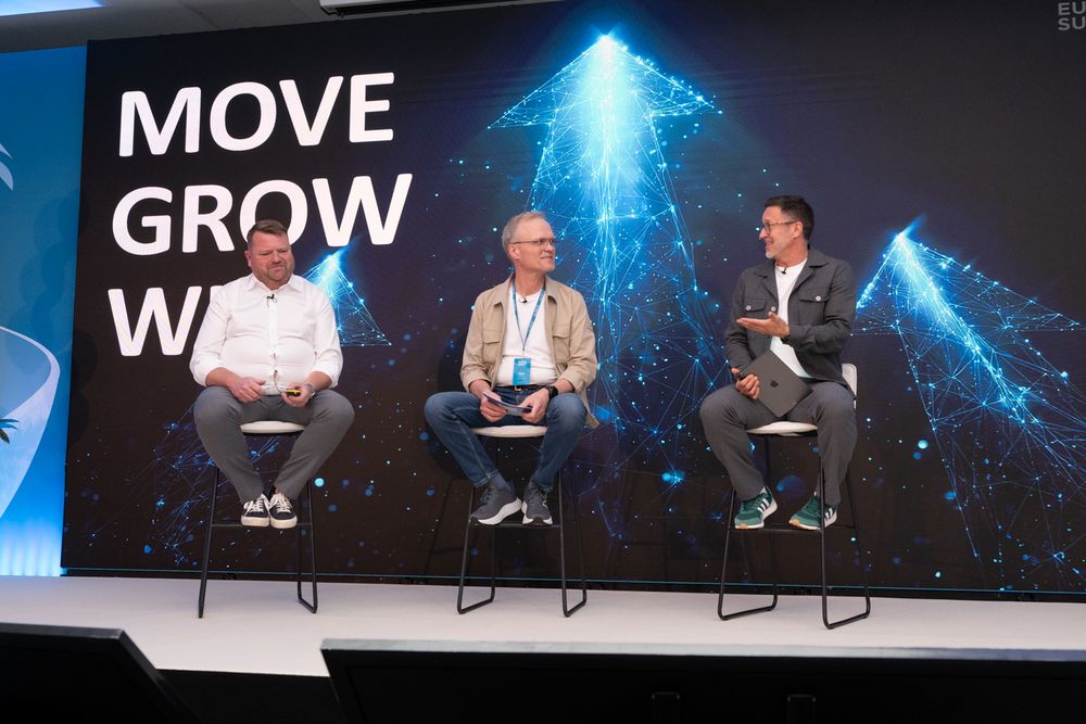 Three speakers participate in a panel discussion on stage with "MOVE GROW WIN" text and blue lighting in the background.