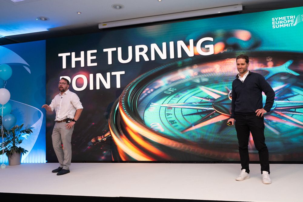 Two presenters stand on stage in front of a large screen displaying "THE TURNING POINT" text with blue lighting.