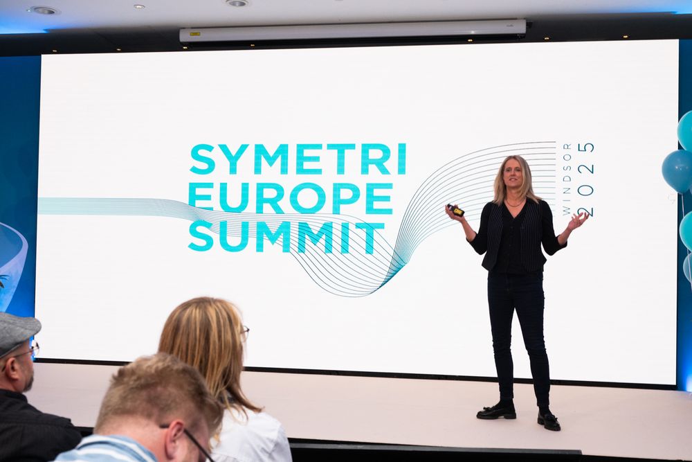 Speaker gestures while presenting on stage at the Symetri Europe Summit in front of a large digital display screen.