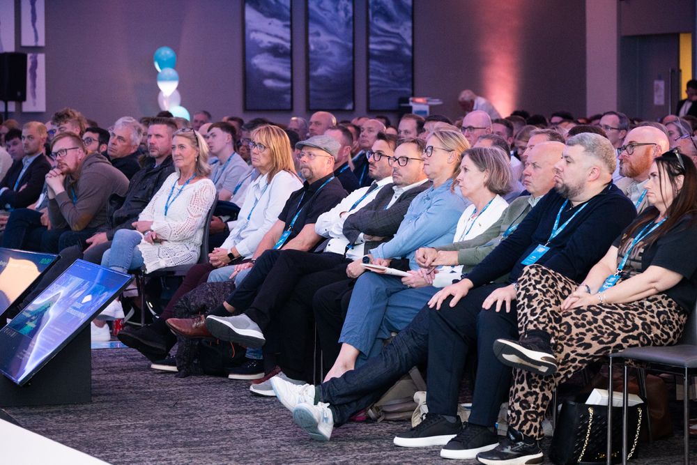 A large audience sits attentively in rows during a conference or presentation event with colored lighting.