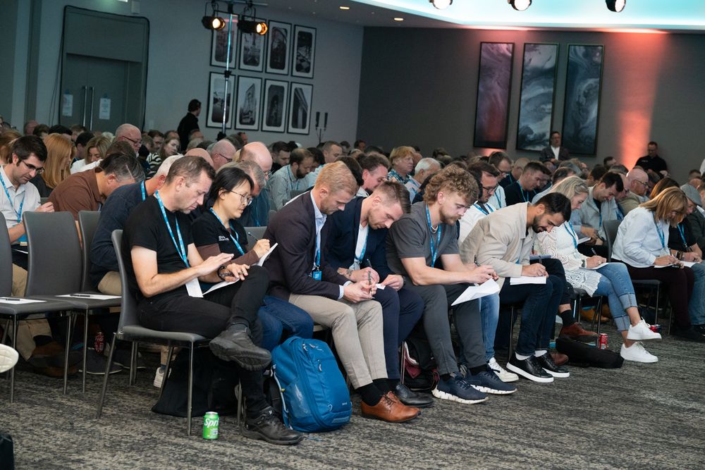 A crowded audience sits in rows looking down at their phones and devices during what appears to be a conference or meeting.