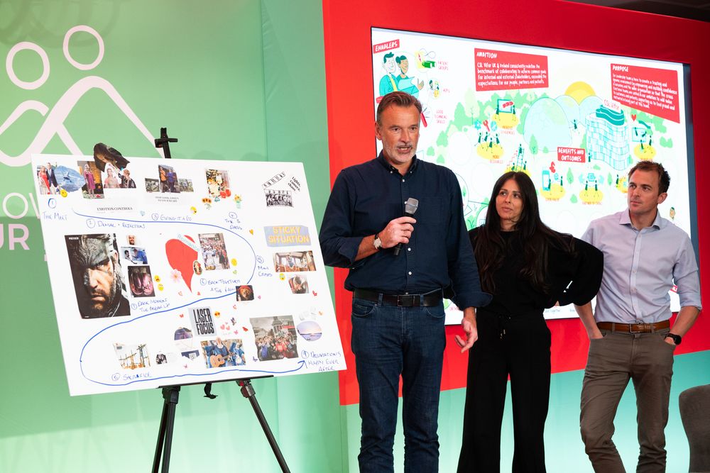 Three presenters stand near a display board with photos and colorful artwork during a presentation event.