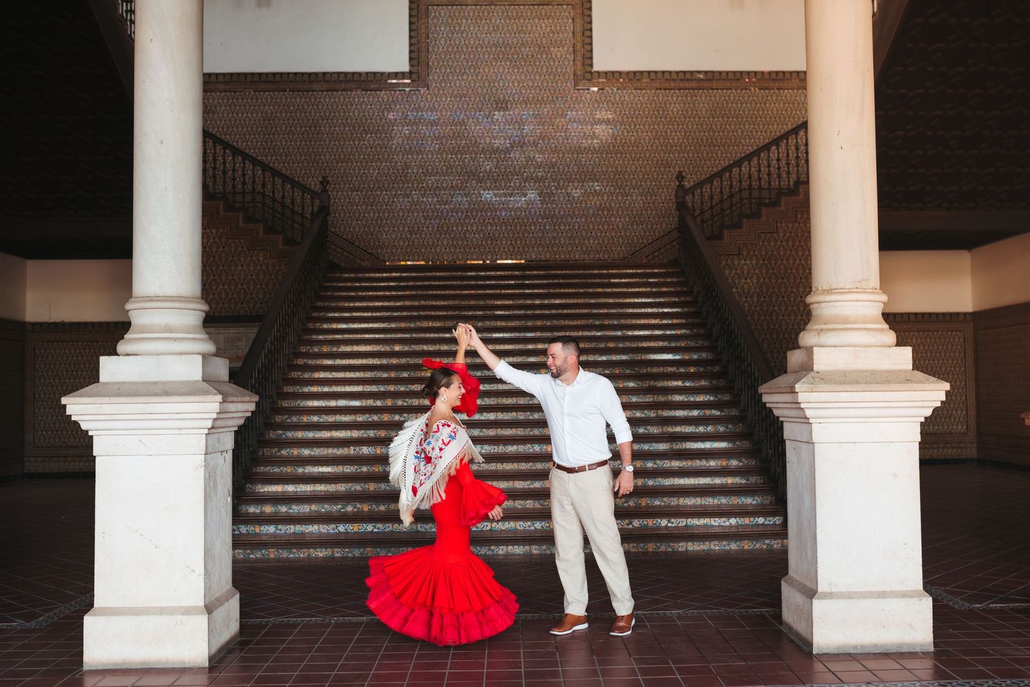 A couple in red and white dance attire poses on a grand staircase with elegant white pillars in an ornate building.
