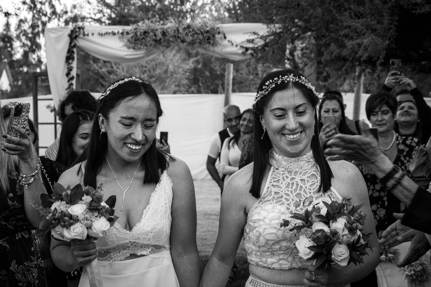 Novias caminando de la mano tras la ceremonia, sonrientes y sosteniendo sus ramos mientras los invitados celebran. Blanco y negro.