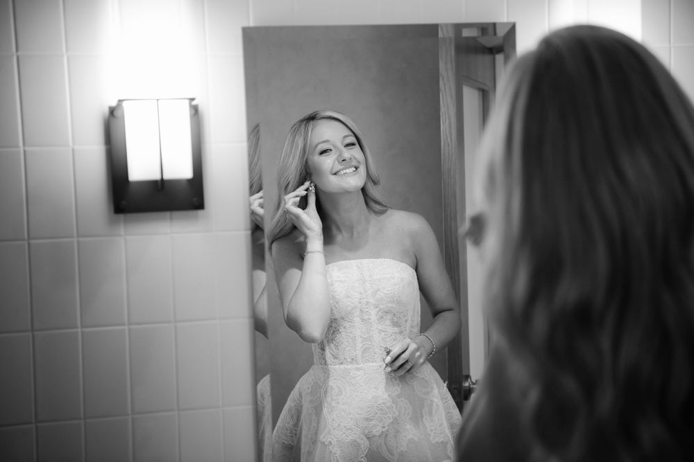 A bride in a strapless white wedding dress applies makeup while looking in a bathroom mirror.