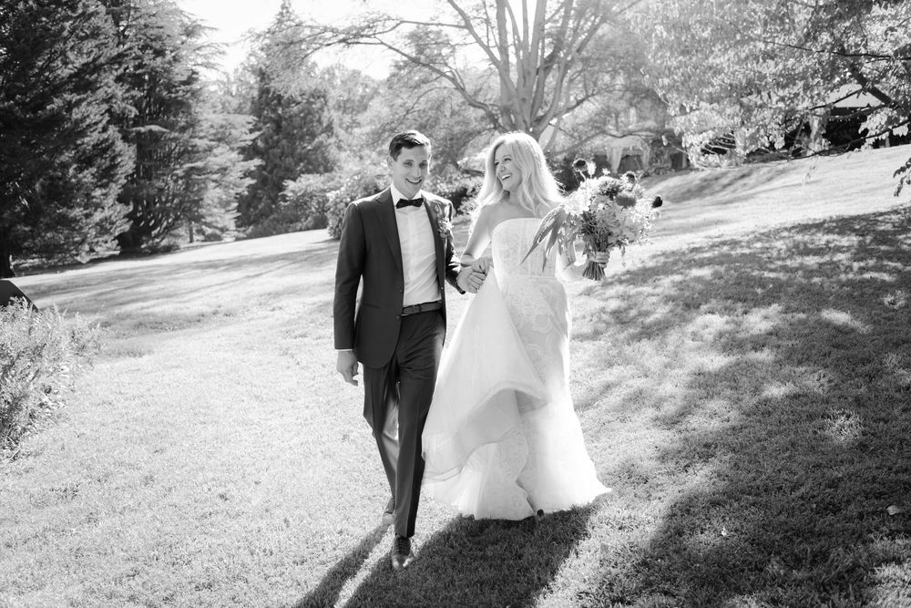 A couple in wedding attire walks together across a sunlit garden lawn during their outdoor wedding celebration.