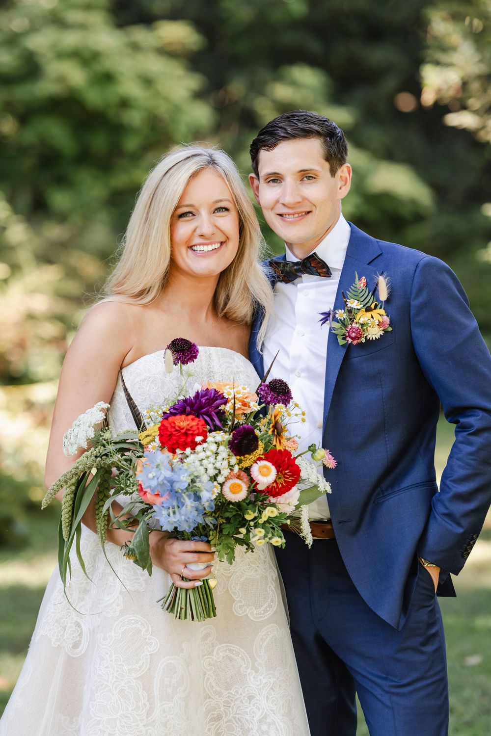A bride in a white lace gown and groom in navy suit pose outdoors with a vibrant autumn bouquet.