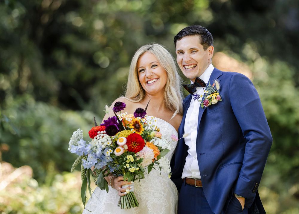A happy couple poses outdoors on their wedding day with a vibrant autumn bouquet and navy suit.