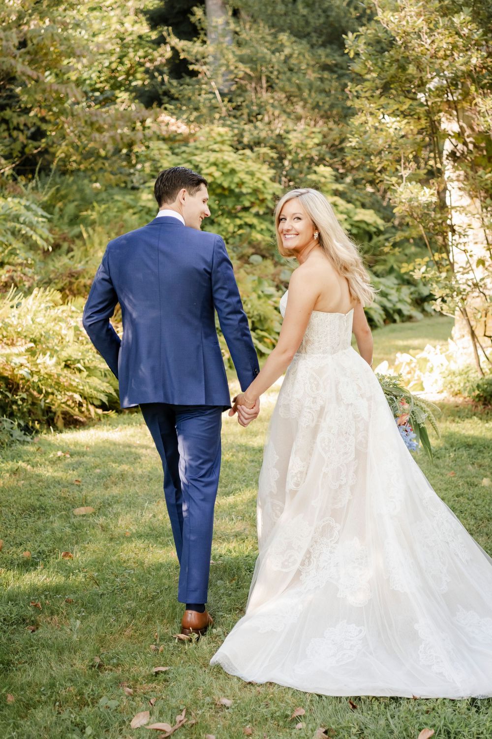 A romantic wedding couple walks together through a sunlit garden path in elegant navy suit and lace gown.
