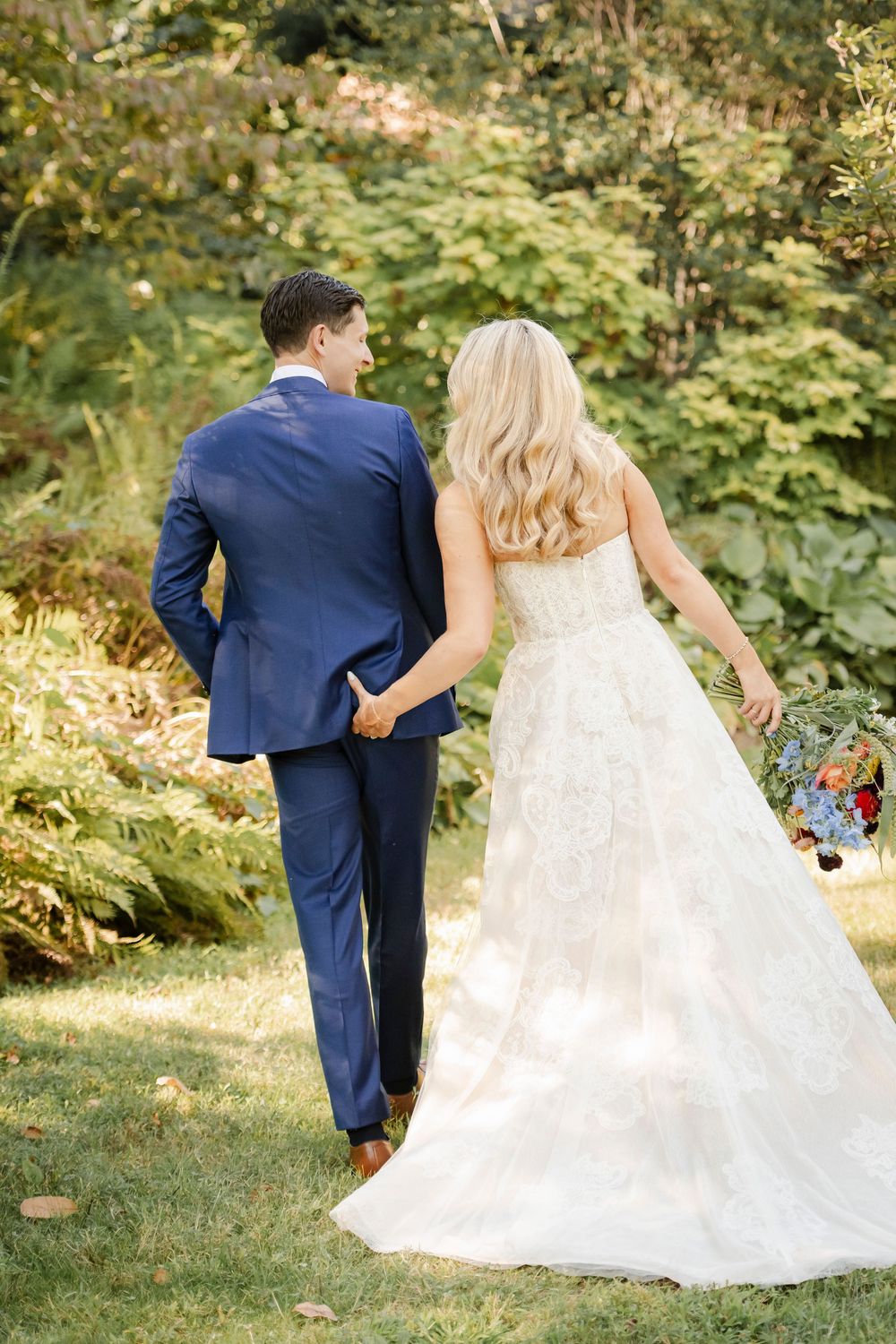 A couple in wedding attire walking together through a lush garden setting with greenery and ferns.