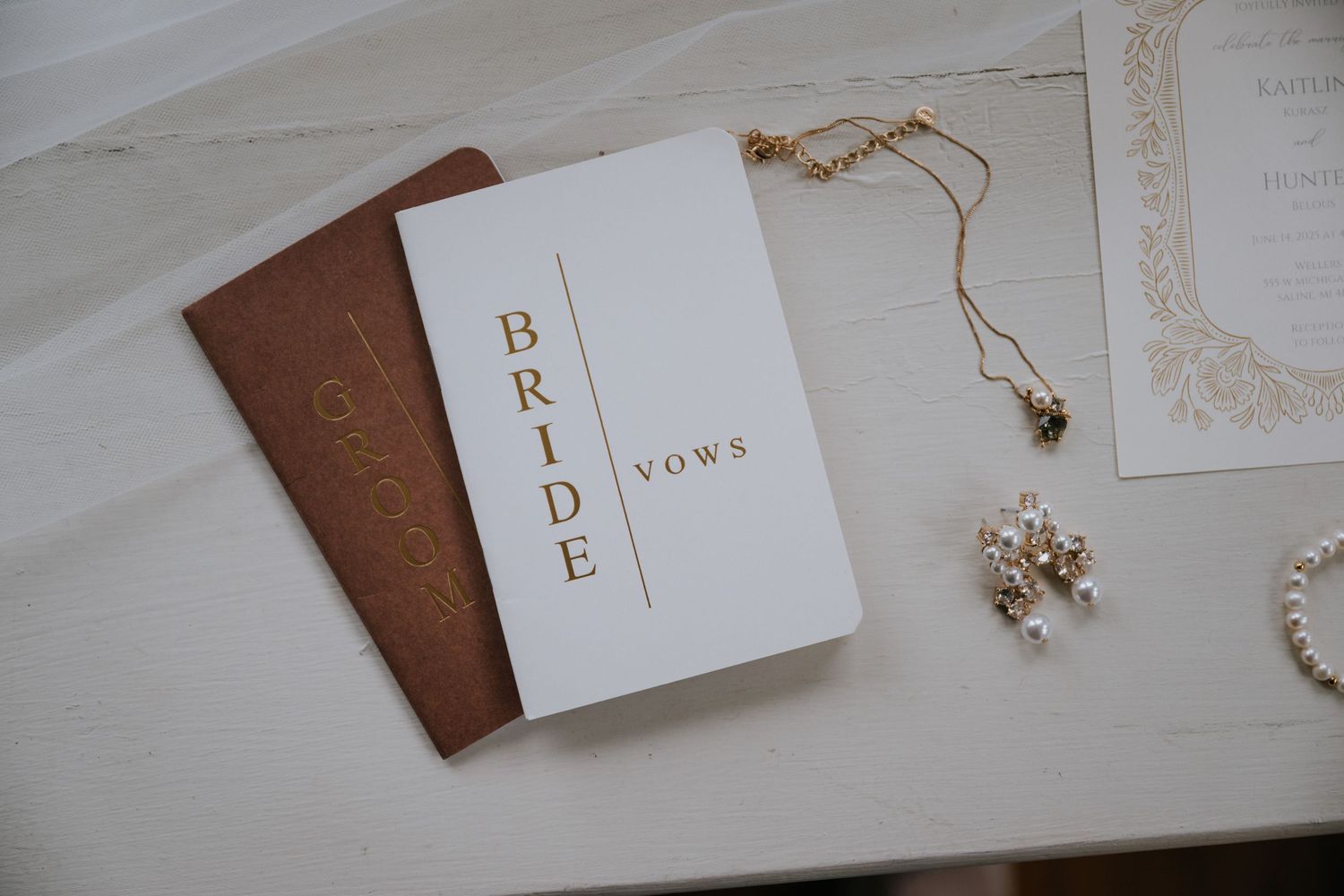 Elegant wedding stationery and accessories including vow books and jewelry displayed on white surface.